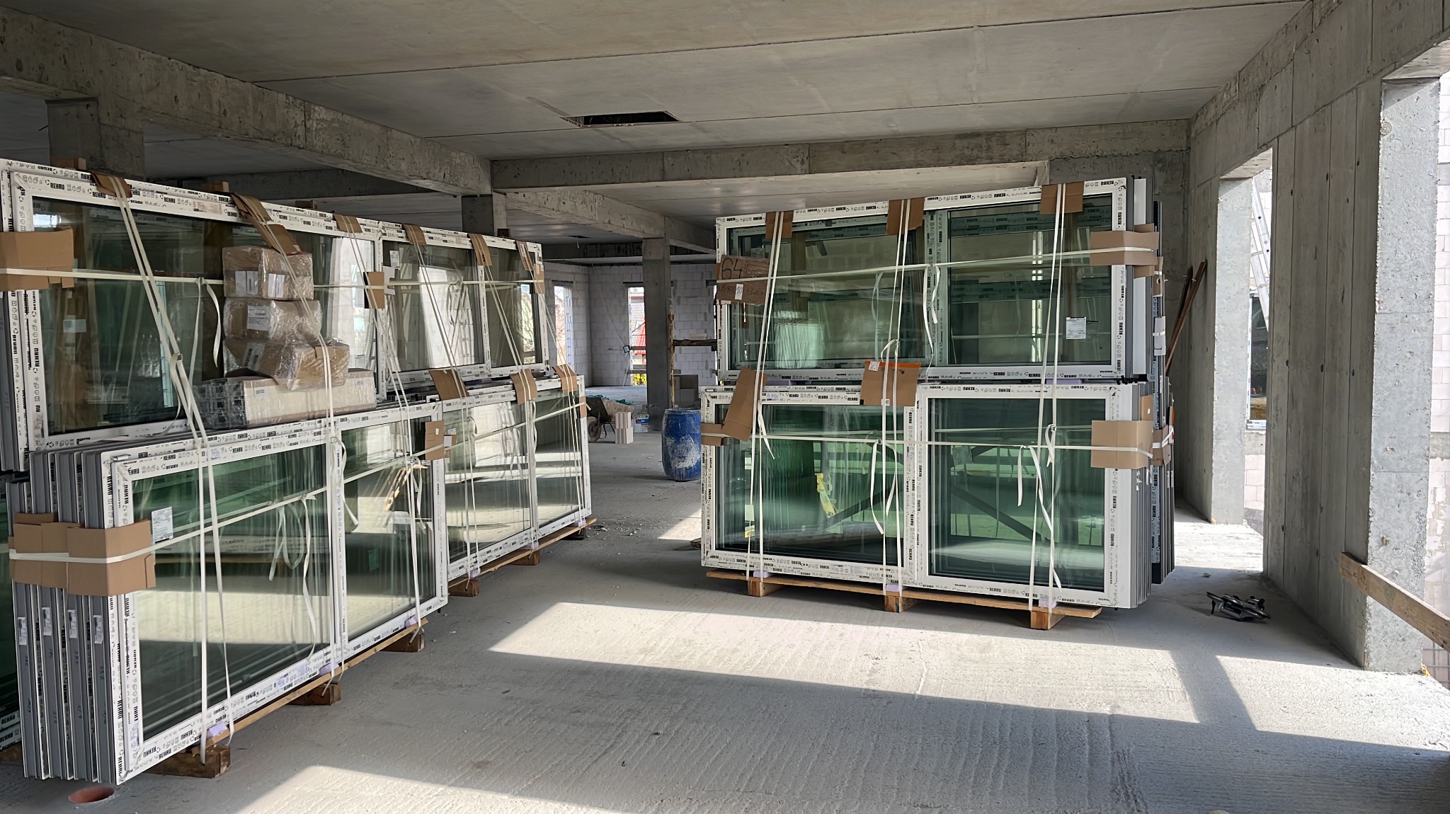 Białe okna PCV, zabezpieczone taśmą, ułożone na paletach w surowym wnętrzu budynku w trakcie budowy. Widoczne betonowe ściany i sufit.