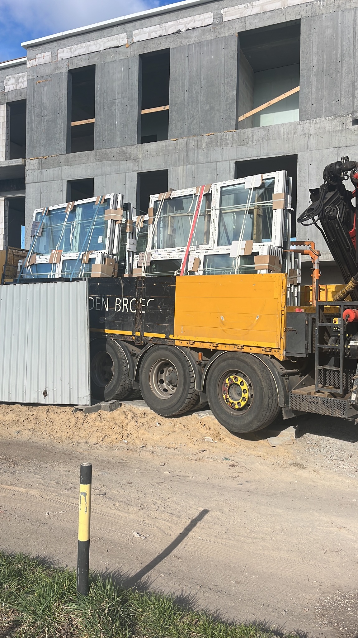Transport okien na budowę. Ciężarówka z ramą HDS przewozi okna PCV na plac budowy, gdzie widać surowy budynek w tle. Żółto-czarna skrzynia ładunkowa.
