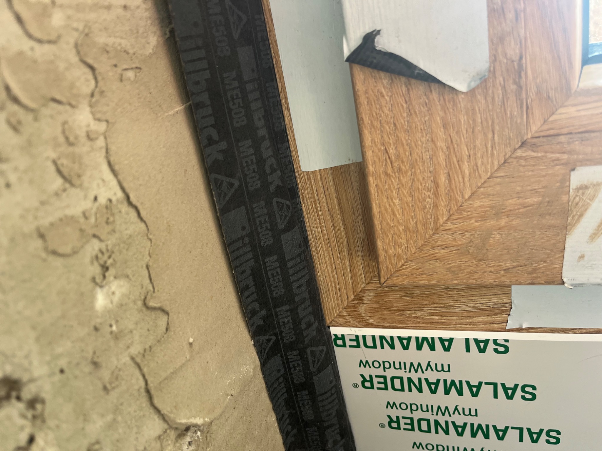 Detal montażu okna Salamander z uszczelnieniem Illbruck ME508 przy surowej ścianie. Widoczny narożnik ramy okiennej o fakturze drewna i taśma uszczelniająca.