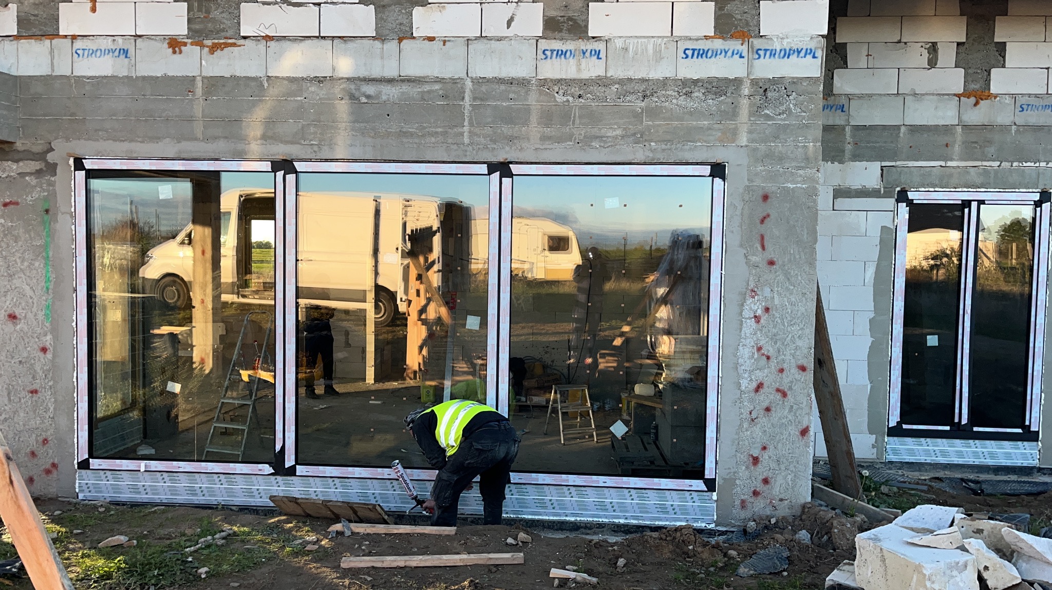 Montaż przesuwnych okien tarasowych w surowym budynku. Widoczny pracownik w kamizelce, narzędzia i elementy konstrukcyjne.