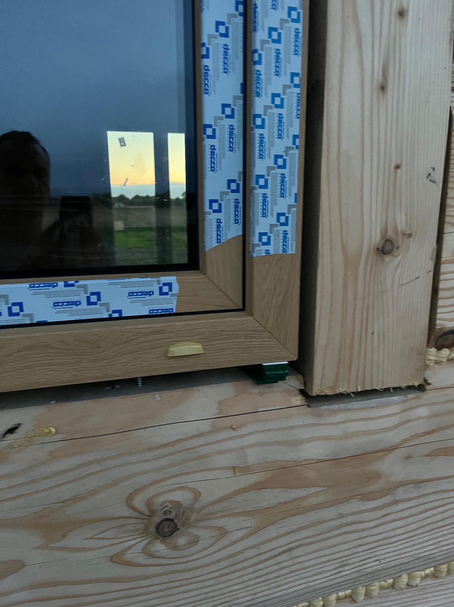 Fragment okna z drewnopodobną ramą, zabezpieczonego folią DECCO, zamontowanego w drewnianej konstrukcji. Widoczne detale montażu i pianka uszczelniająca.