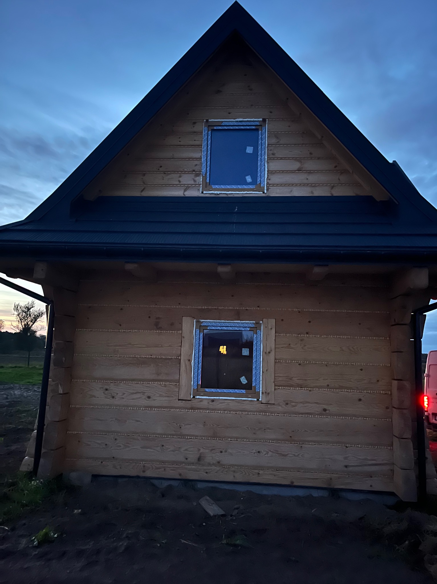 Dom z bali w trakcie montażu okien. Widoczne okna z folią ochronną, drewniana konstrukcja i czarny dach. Ujęcie z dołu, wieczorne światło.