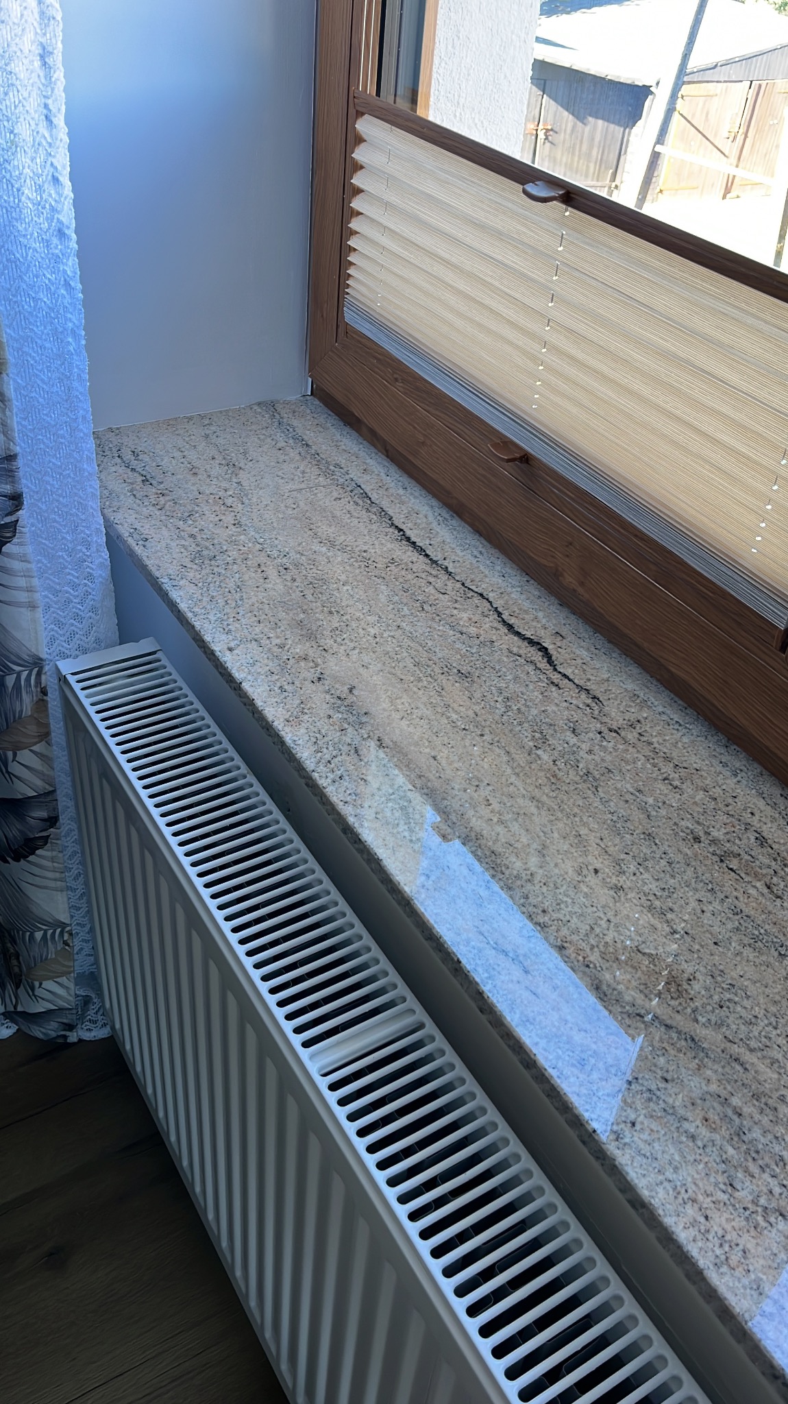 Beżowy parapet z kamienia, brązowe okno z plisowaną roletą i biały grzejnik. Ujęcie z góry, widoczny fragment podłogi i zasłony w tle.