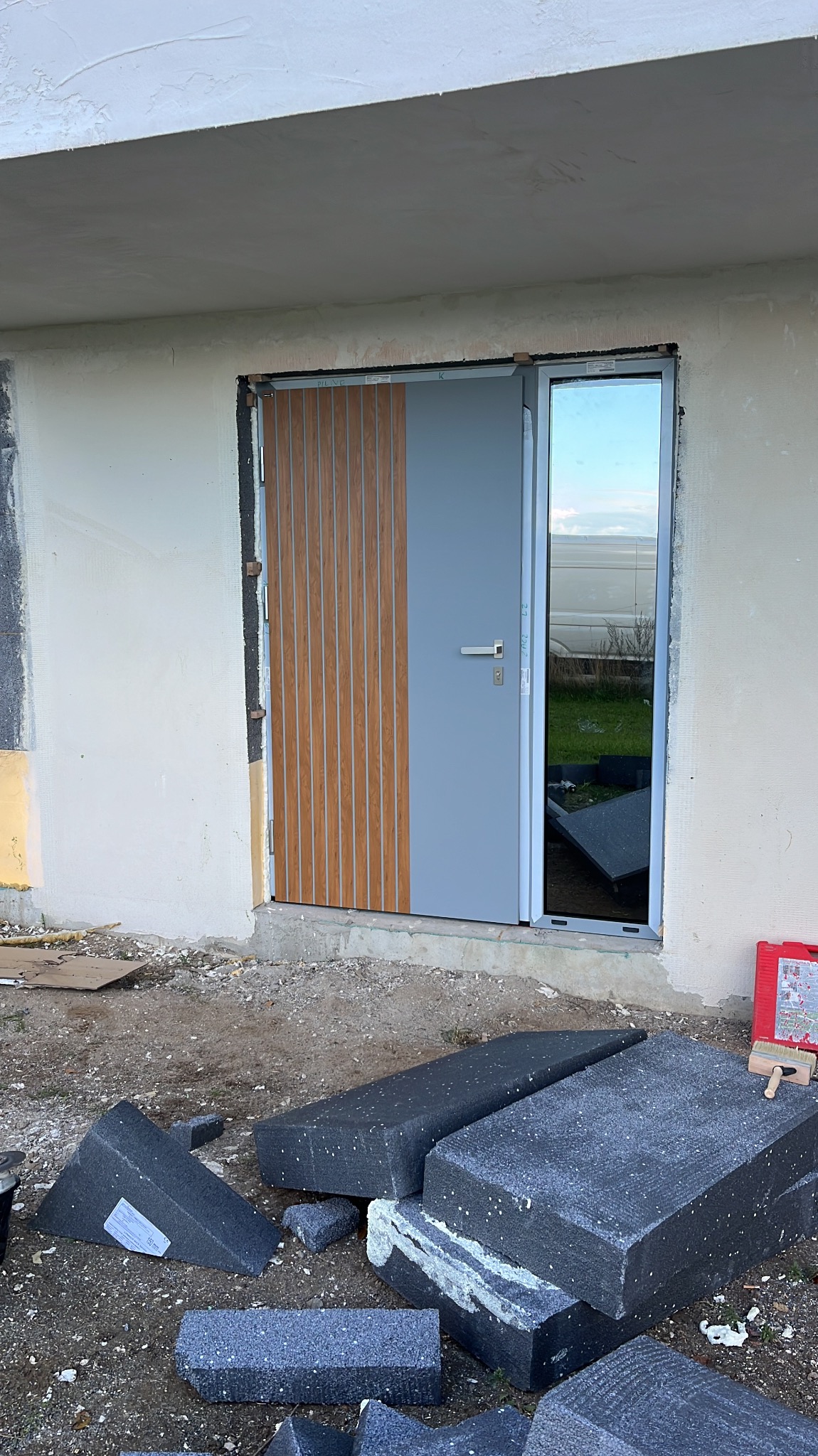 Nowe, szare drzwi zewnętrzne z drewnianymi panelami, zamontowane w surowym otworze budynku. Fragmenty styropianu na ziemi. Widoczne odblaski w szybie.