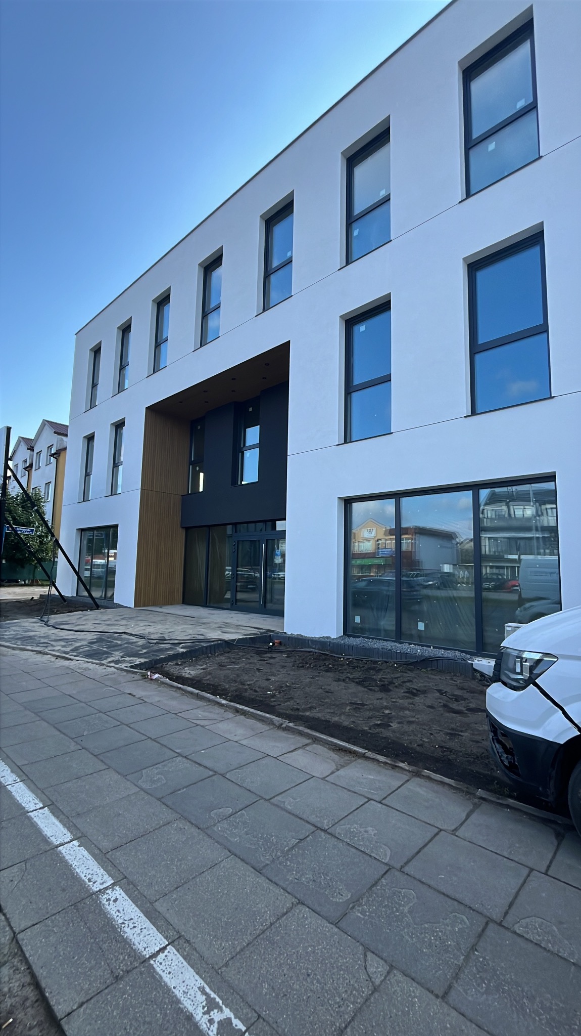 Nowoczesny budynek biurowy z białymi ścianami i dużymi oknami z czarnymi ramami. Widoczne elementy drewniane i fragment samochodu.