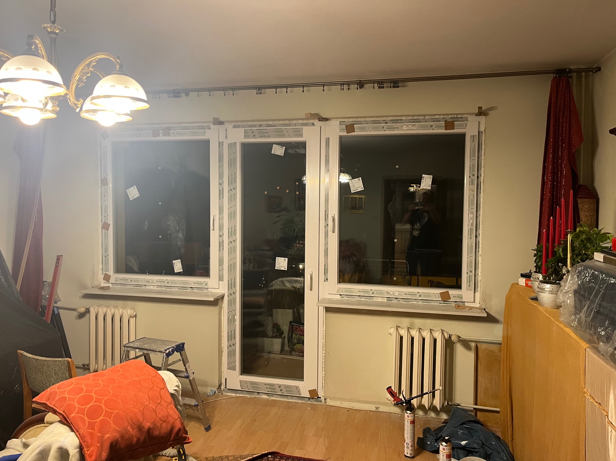Montaż okien PCV w mieszkaniu: widoczne okna Salamander z folią ochronną, pianka montażowa, drabina i grzejnik. Wnętrze w trakcie prac wykończeniowych.