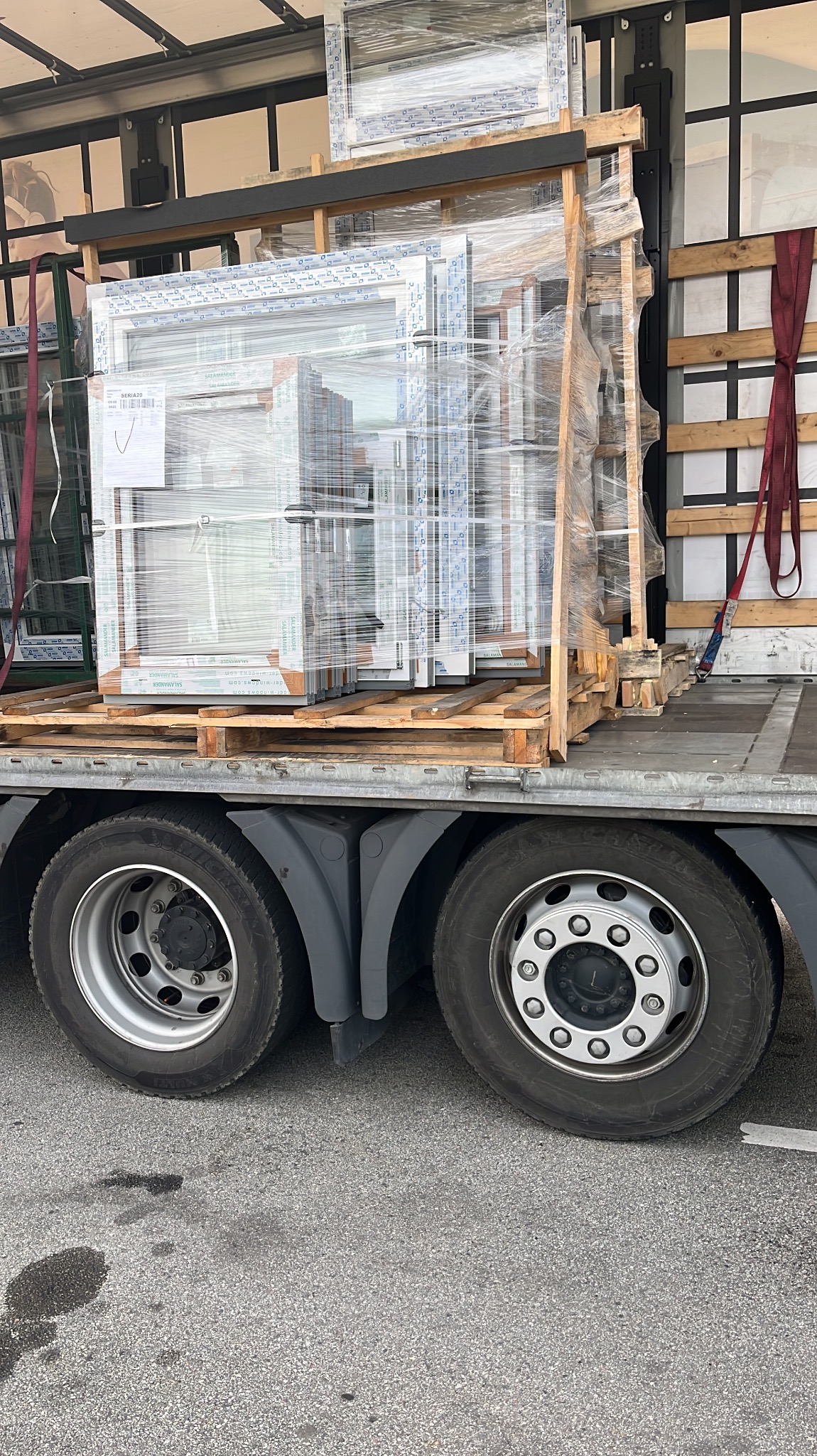Transport okien PCV Salamander na palecie, zabezpieczonych folią stretch, widoczne koła ciężarówki, etykieta z informacjami o produkcie.