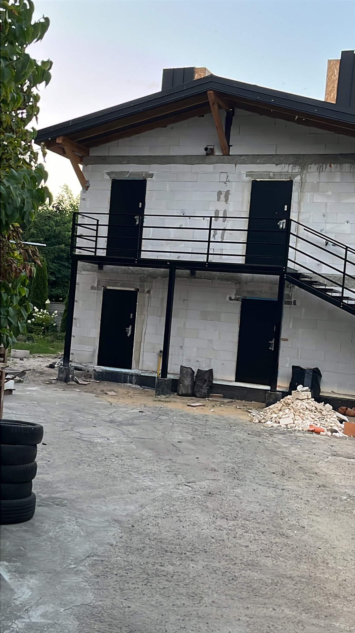 Surowy budynek z białego bloczka z czarnymi drzwiami, balkonem i schodami. Widoczne są ślady prac budowlanych i opony samochodowe.