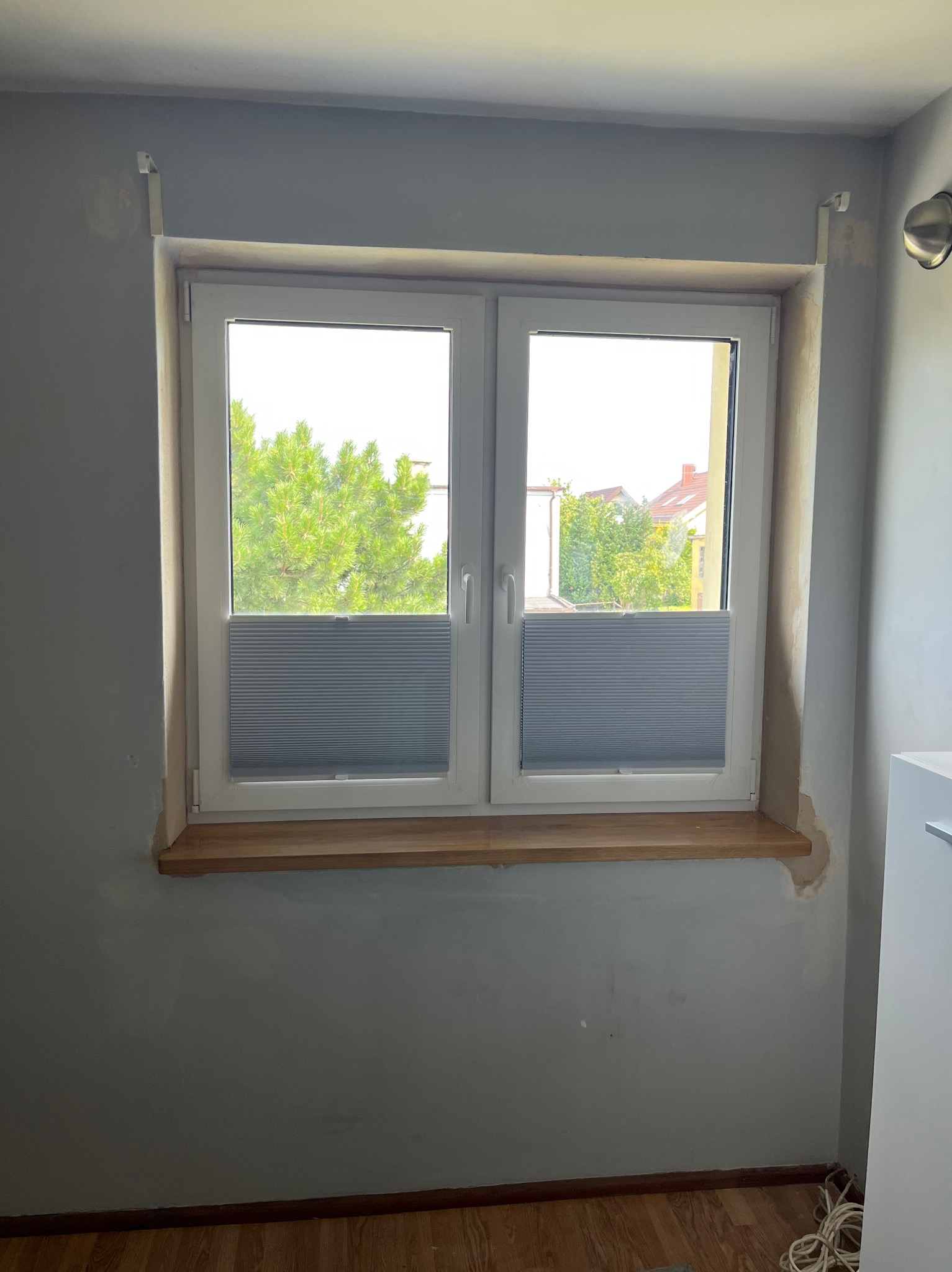 Białe okno PCV z zamontowaną szarą plisą okienną, drewniany parapet, szare ściany w trakcie wykańczania, widok na zieleń za oknem.
