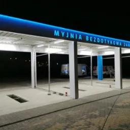 Konstrukcja stalowa z niebieskim oświetleniem LED nad wejściem do bezdotykowej myjni samochodowej 24h, widoczne kratki odpływowe w posadzce i elementy montażowe.