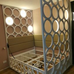 ZEKO STAL ZBIGNIEW MALIK - Biała, metalowa konstrukcja łóżka z ażurowym wezgłowiem i bokami, zdobionymi okręgami, z podświetleniem LED, na drewnianej podłodze, z beżowym tapicerowanym zagłówkiem.