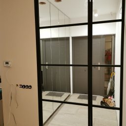 ZEKO STAL ZBIGNIEW MALIK - Szklana ścianka działowa z czarnymi profilami, widoczne niedokończone prace elektryczne na ścianie obok, odbicie dwóch szarych drzwi w szkle.