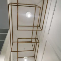 Geometryczna konstrukcja z metalowych profili w kolorze złotym, tworząca przestrzenne ramy na suficie podwieszanym, z widocznymi dwoma lampami wiszącymi o białych kloszach.