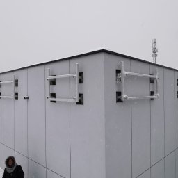 ZEKO STAL ZBIGNIEW MALIK - Szare elementy konstrukcyjne, prawdopodobnie wsporniki, przymocowane do elewacji budynku w trakcie śnieżnej pogody. Część elementów zabezpieczona folią. W tle widoczna wieża telekomunikacyjna...