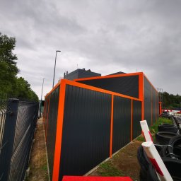 ZEKO STAL ZBIGNIEW MALIK - Nowoczesne ogrodzenie panelowe w kolorze grafitowym, z pomarańczowymi akcentami, otaczające tor kartingowy z oponami zabezpieczającymi, widoczne pod pochmurnym niebem.