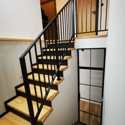 ZEKO STAL ZBIGNIEW MALIK - Drewniane schody z czarną, metalową balustradą i czarnymi podstopnicami, widziane z góry, z przeszklonymi drzwiami w tle.