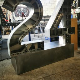 ZEKO STAL ZBIGNIEW MALIK - Duże, metalowe cyfry '24' w trakcie spawania, widoczne spawy punktowe, warsztatowe otoczenie z maszyną do obróbki metalu w tle i banerem z motywem czaszek.