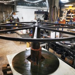 ZEKO STAL ZBIGNIEW MALIK - Konstrukcja metalowa w kształcie koła z centralnym wspornikiem, prawdopodobnie element większej instalacji, w warsztacie z widocznym sprzętem spawalniczym i narzędziami.