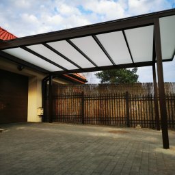 ZEKO STAL ZBIGNIEW MALIK - Zadaszenie nad miejscem parkingowym przy domu jednorodzinnym, konstrukcja z brązowych profili i białych paneli dachowych, widoczny fragment bramy garażowej, ogrodzenia i kostki brukowej.