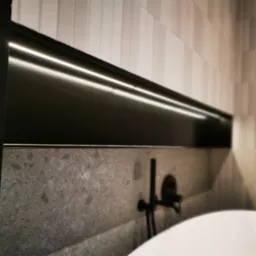 Minimalistyczna łazienka z podświetlaną półką nad wanną, ściany wykończone płytkami w odcieniach szarości, czarna bateria wannowa na tle kamiennej ściany.