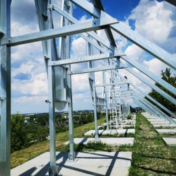ZEKO STAL ZBIGNIEW MALIK - Metalowa konstrukcja wsporcza z drabinką, prawdopodobnie pod panele słoneczne, na betonowych fundamentach, widok z dołu, w tle zielony krajobraz i błękitne niebo z chmurami.