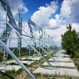 ZEKO STAL ZBIGNIEW MALIK - Konstrukcja metalowa z drabinką na betonowych fundamentach na wzgórzu, widok na miasto w tle pod błękitnym niebem z chmurami.