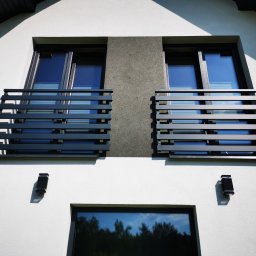 ZEKO STAL ZBIGNIEW MALIK - Dwa balkony z nowoczesnymi, poziomymi balustradami w kolorze grafitowym, zamontowane przy oknach na białej fasadzie domu z szarą wstawką. Widoczne czarne lampy zewnętrzne i odbicie drzew w dolnym...