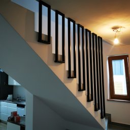 ZEKO STAL ZBIGNIEW MALIK - Nowoczesne schody z białą konstrukcją i czarnymi, pionowymi elementami dekoracyjnymi, widoczne z dołu, z niedokończonym oświetleniem sufitowym i oknem w tle.