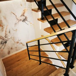 ZEKO STAL ZBIGNIEW MALIK - Drewniane schody z czarną metalową balustradą i drewnianymi poręczami, na ścianie dekoracyjny motyw żurawi.