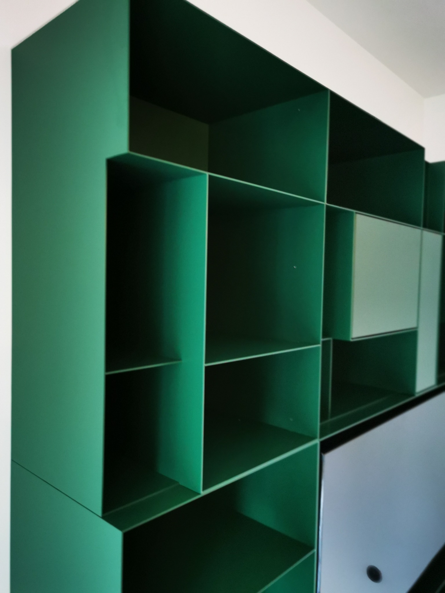 Zielony, metalowy regał o geometrycznym kształcie z otwartymi półkami i kilkoma zamykanymi szafkami z jasnymi frontami.