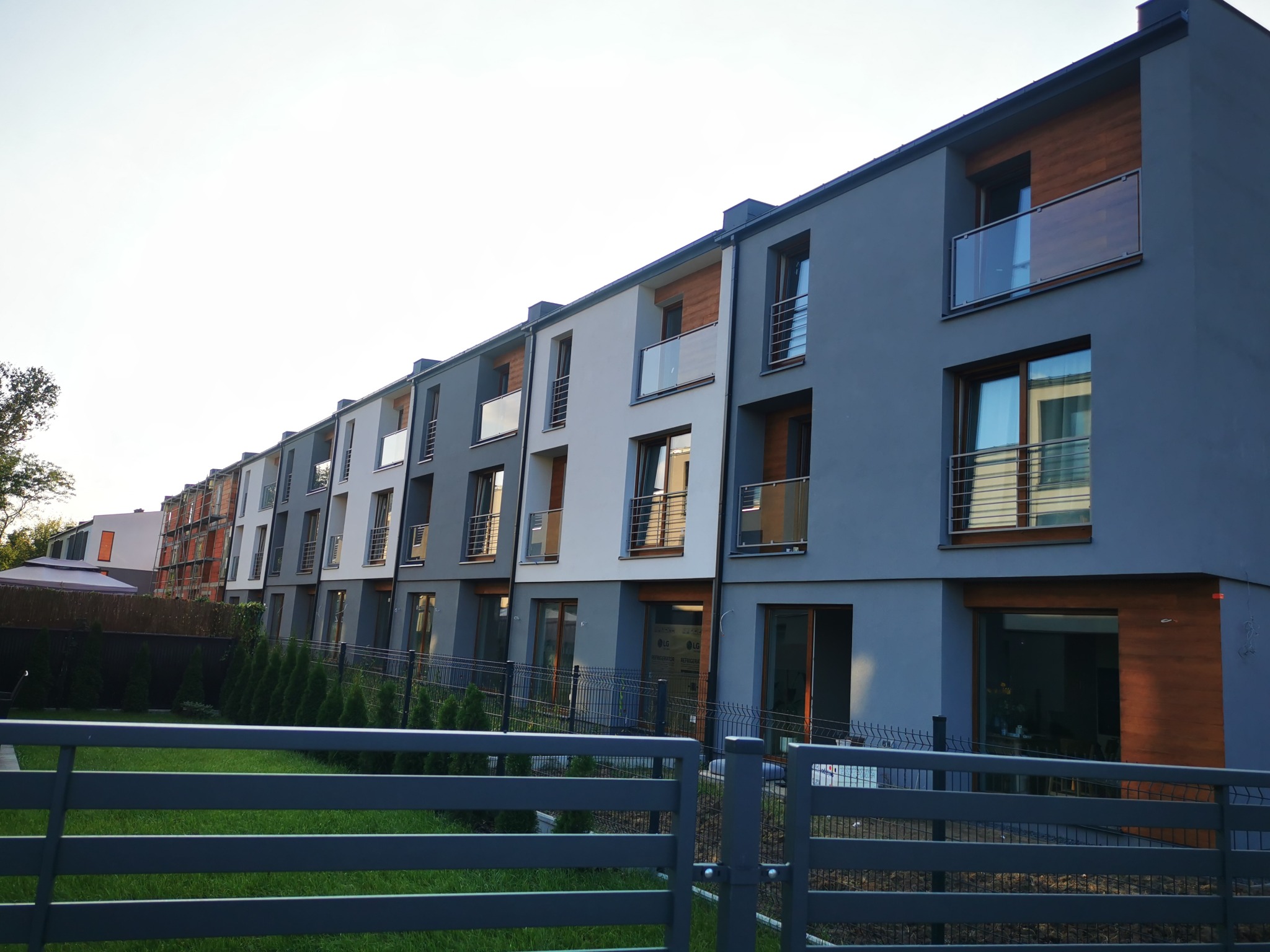 Szeregowiec z balkonami z balustradami szklanymi, widok zza metalowego ogrodzenia panelowego.