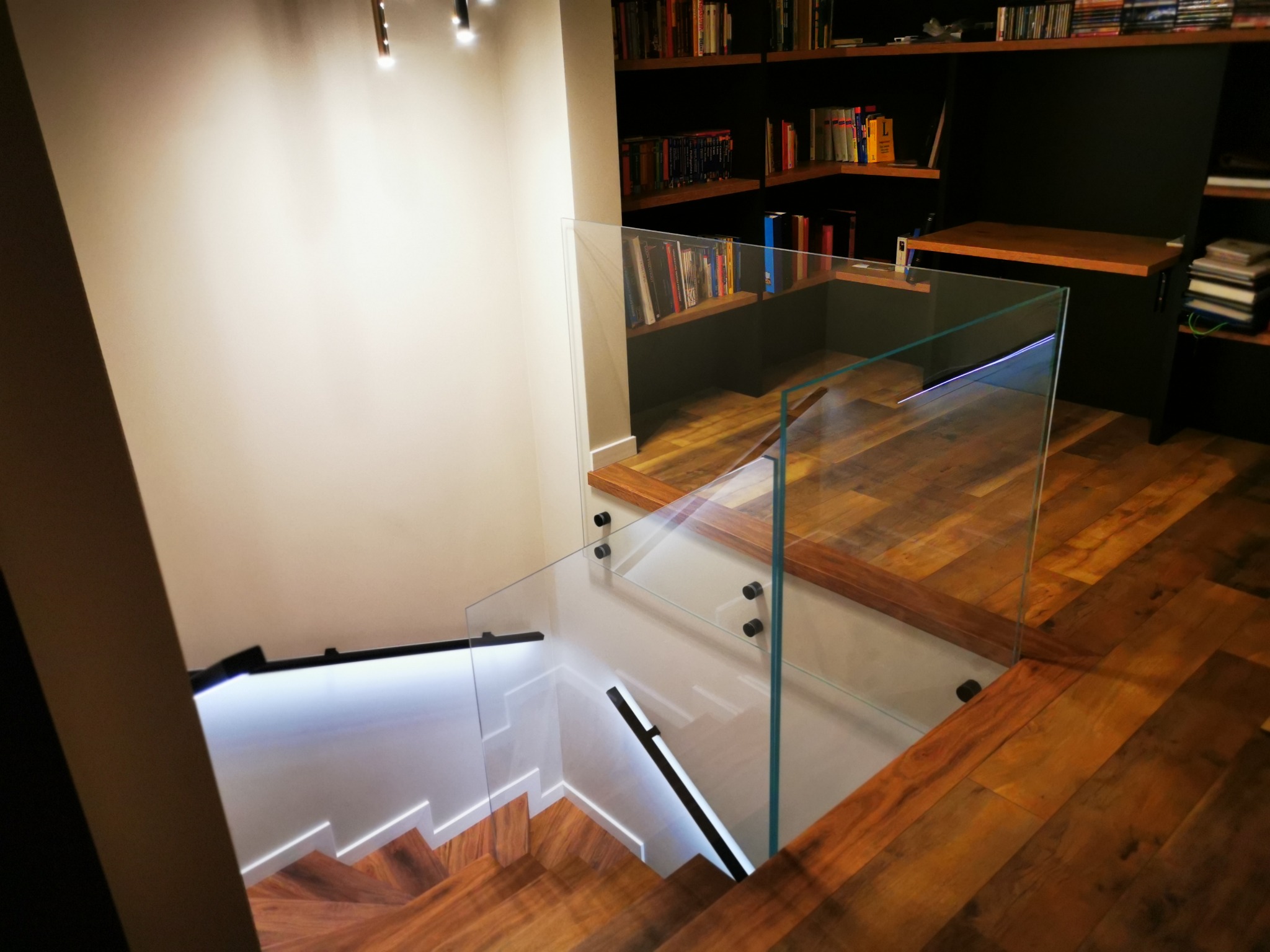 Nowoczesne schody z drewnianymi stopniami i szklaną balustradą mocowaną punktowo, podświetlone listwą LED, widok z góry, w tle regał z książkami.