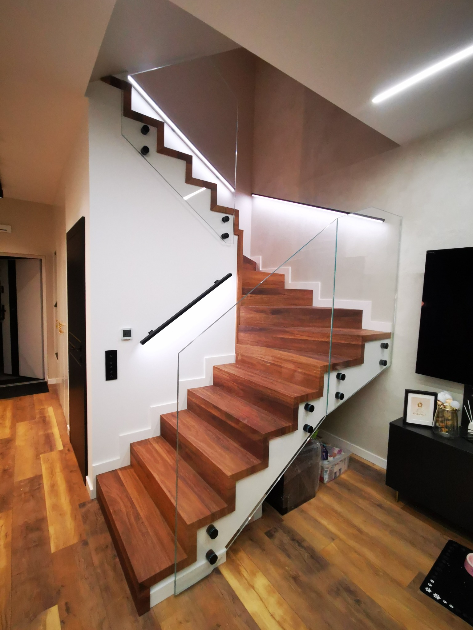 Nowoczesne schody drewniane z balustradą szklaną, podświetlane od spodu, z czarnymi elementami mocującymi szkło i poręczą, widoczne z boku w jasnym wnętrzu.