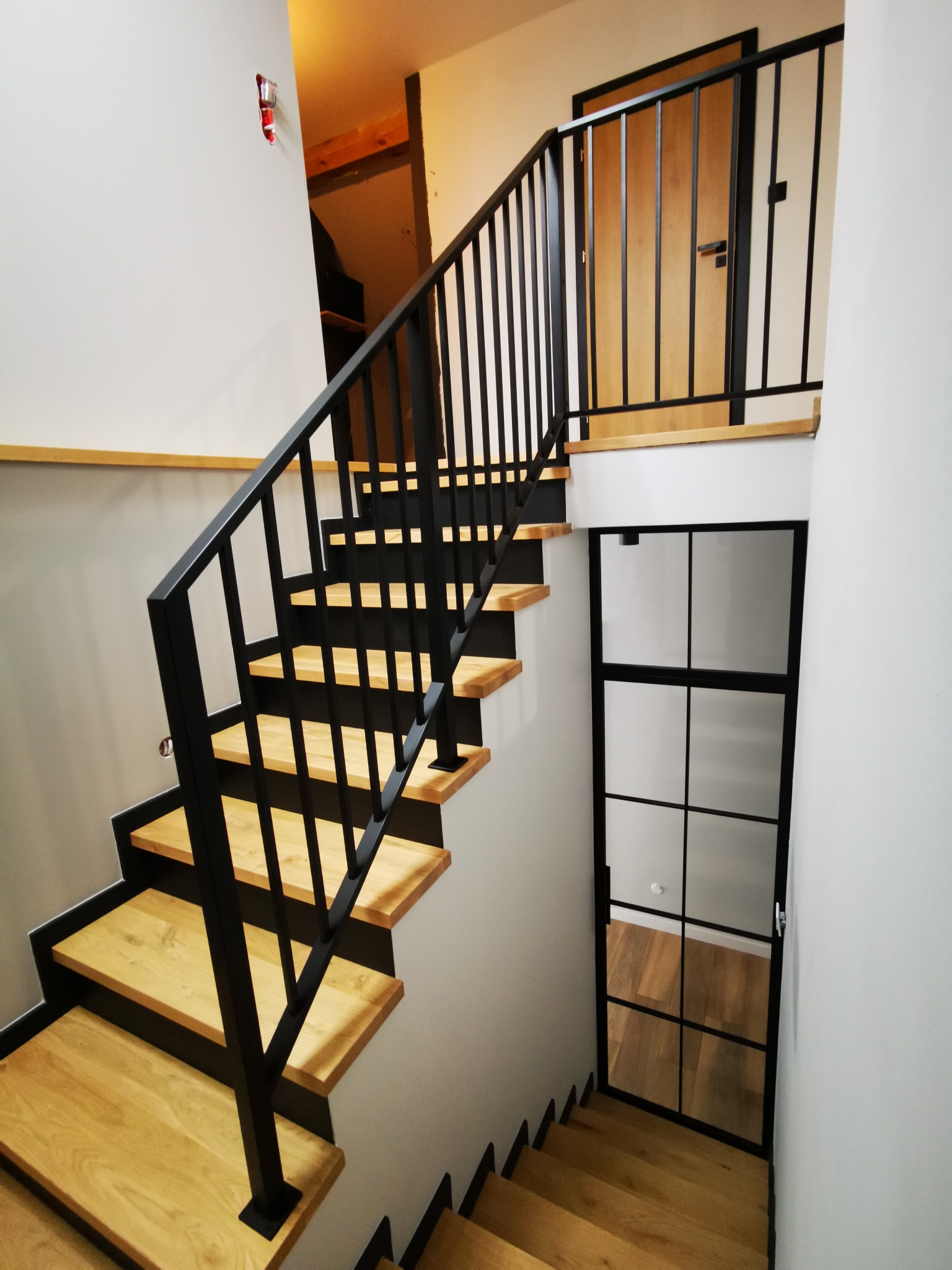 Drewniane schody z czarną, metalową balustradą i czarnymi podstopnicami, widziane z góry, z przeszklonymi drzwiami w tle.