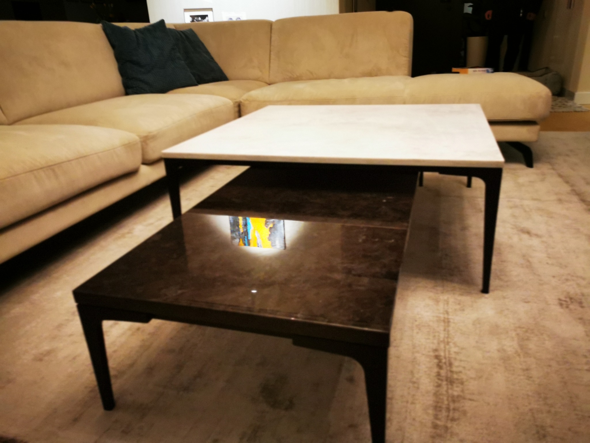 Dwa stoliki kawowe o różnych wysokościach i blatach: górny z białego marmuru, dolny z ciemnego drewna, ustawione na beżowym dywanie w salonie z narożną sofą w tle.