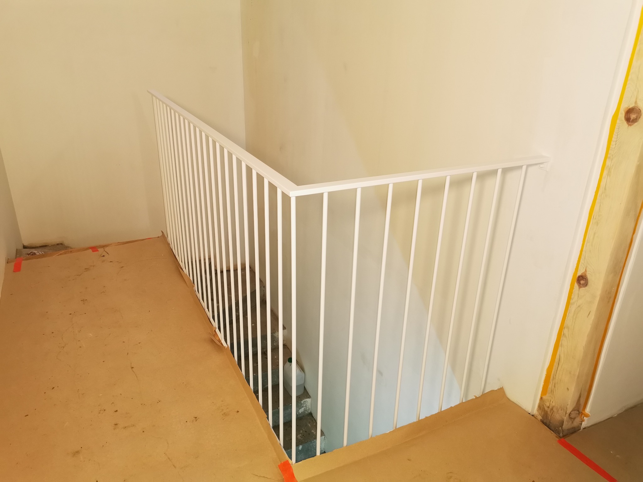 Biała, metalowa balustrada z pionowymi prętami, zamontowana przy schodach w trakcie remontu, z zabezpieczoną podłogą i widocznym fragmentem drewnianej konstrukcji ściany.