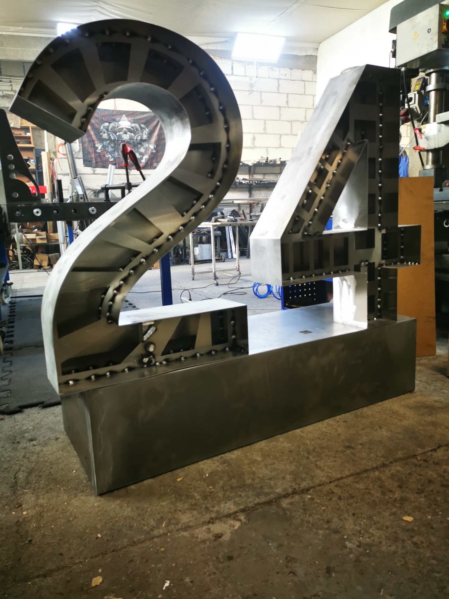 Duże, metalowe cyfry '24' w trakcie spawania, widoczne spawy punktowe, warsztatowe otoczenie z maszyną do obróbki metalu w tle i banerem z motywem czaszek.