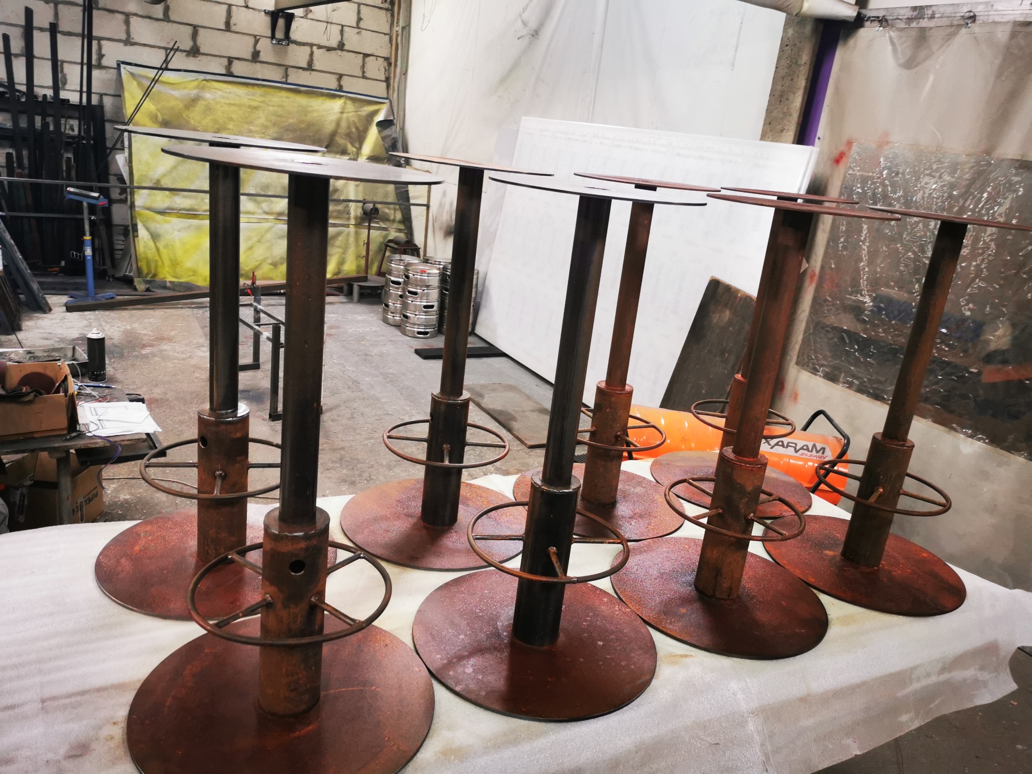 Sześć metalowych stołków barowych z okrągłymi podstawami i siedziskami, pokrytych rdzawym nalotem, ustawionych na białym płótnie w warsztacie.