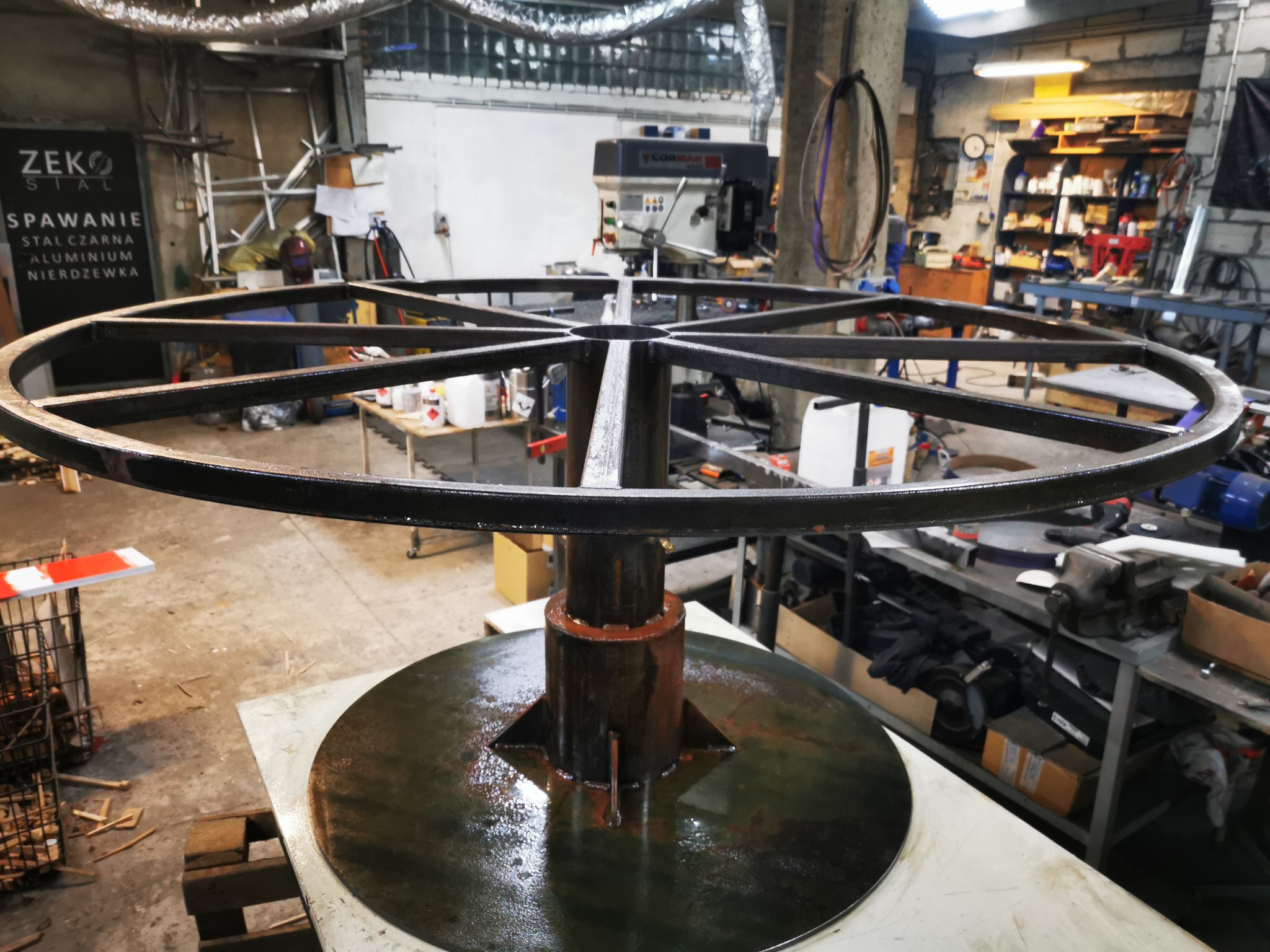 Konstrukcja metalowa w kształcie koła z centralnym wspornikiem, prawdopodobnie element większej instalacji, w warsztacie z widocznym sprzętem spawalniczym i narzędziami.