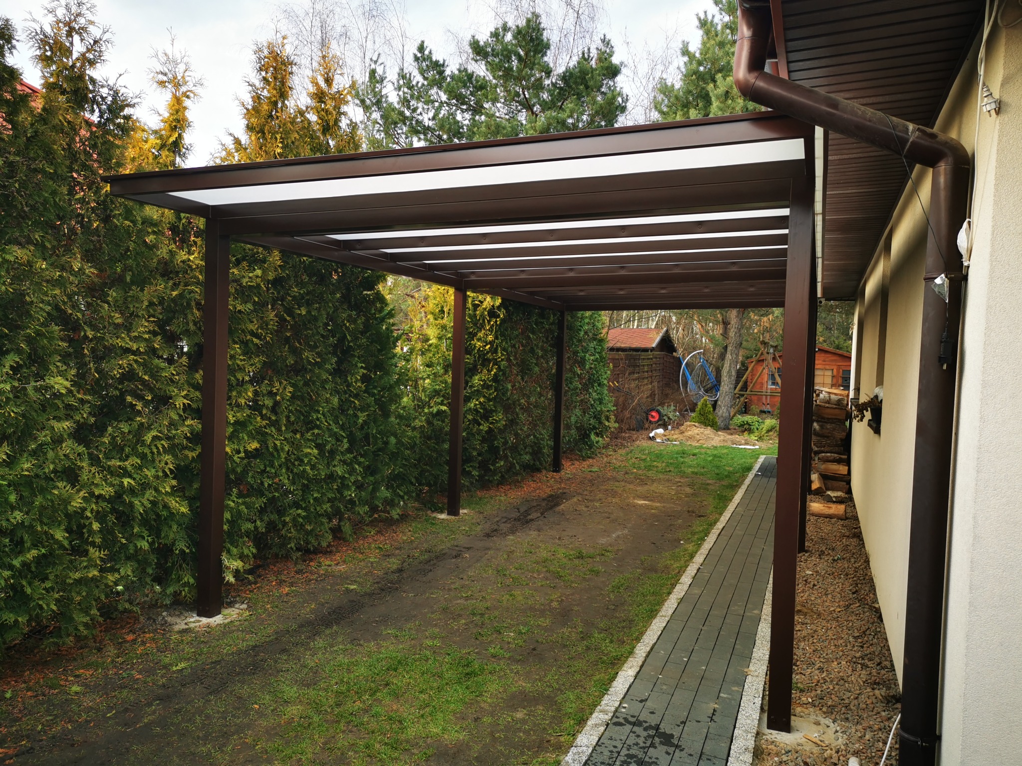 Zadaszenie tarasowe z brązową konstrukcją metalową i białym wypełnieniem, przylegające do ściany budynku, z widocznymi rynnami, otoczone zielenią.