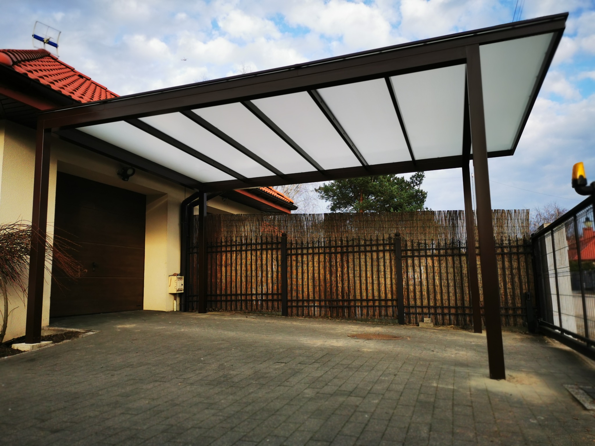 Zadaszenie nad miejscem parkingowym przy domu jednorodzinnym, konstrukcja z brązowych profili i białych paneli dachowych, widoczny fragment bramy garażowej, ogrodzenia i kostki brukowej.