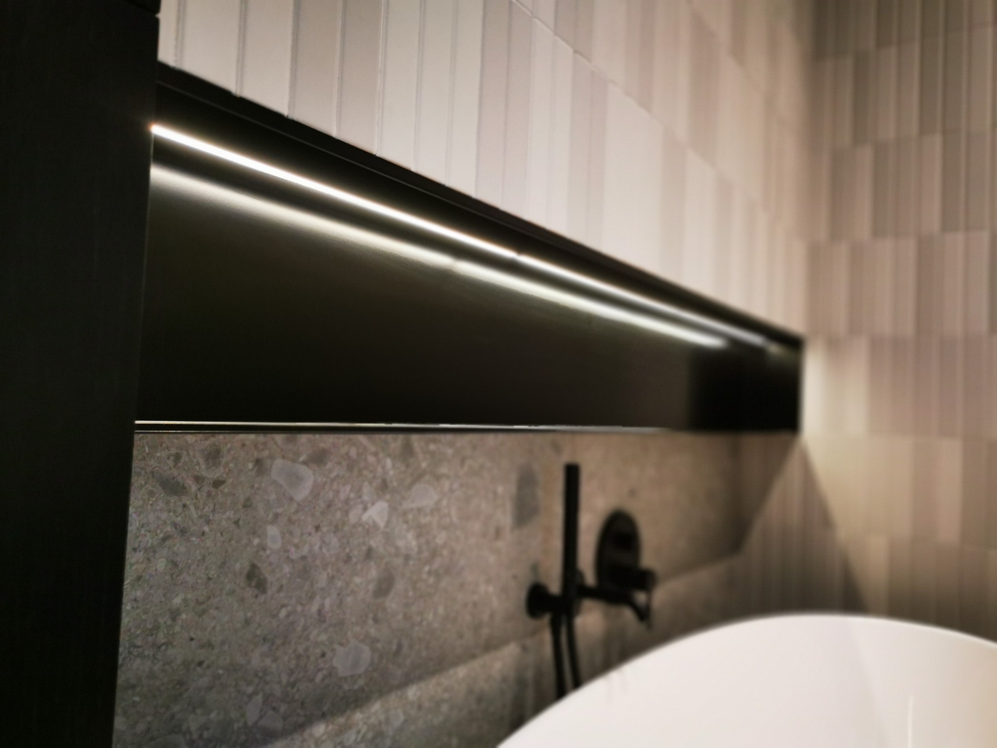 Minimalistyczna łazienka z podświetlaną półką nad wanną, ściany wykończone płytkami w odcieniach szarości, czarna bateria wannowa na tle kamiennej ściany.