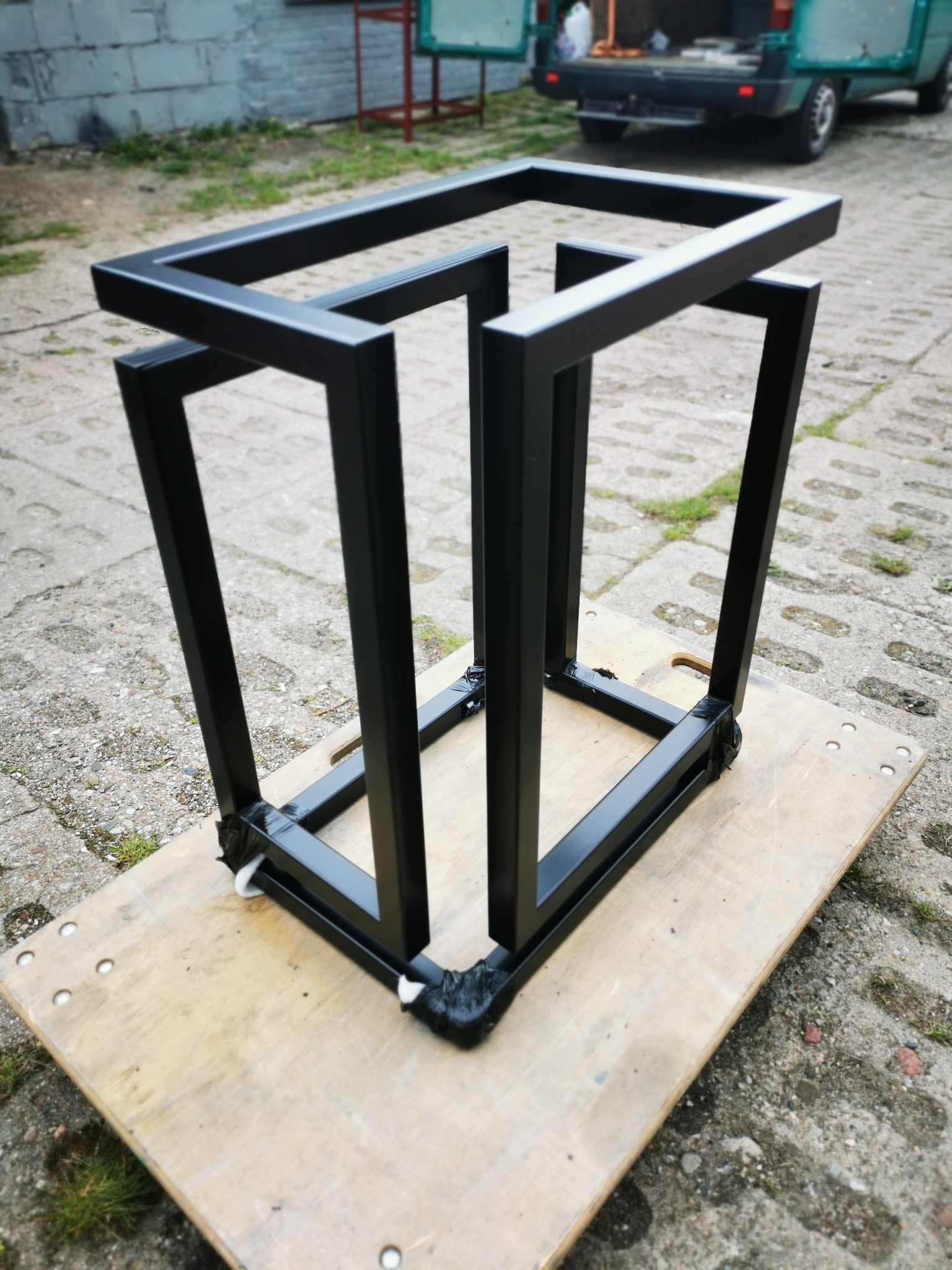 Czarna, metalowa konstrukcja spawana z profili kwadratowych, zabezpieczona taśmą na rogach, umieszczona na płycie OSB na zewnątrz, z widocznym fragmentem samochodu dostawczego w tle.