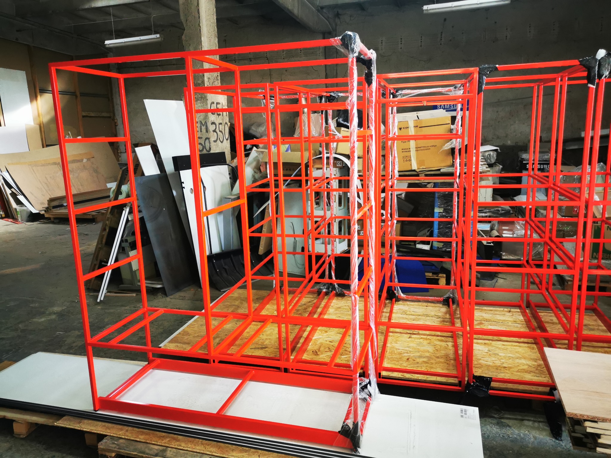 Czerwone, metalowe ramy konstrukcyjne w warsztacie, częściowo owinięte folią ochronną, stojące na paletach, z widocznymi elementami magazynowymi w tle.