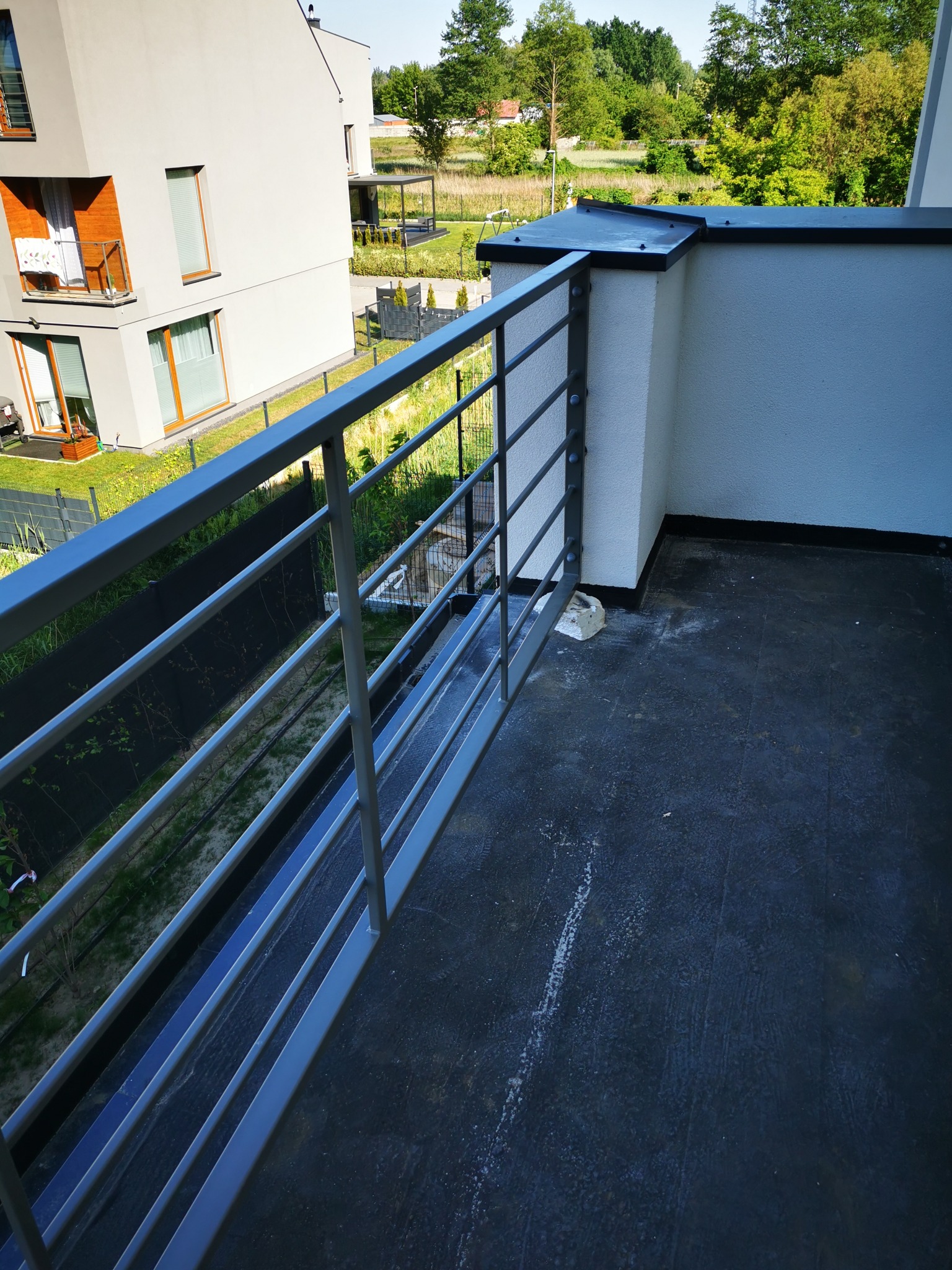 Nowoczesna balustrada balkonowa z poziomych, stalowych profili w Legionowie, widok z góry na czarną, gumową nawierzchnię balkonu i fragment budynku mieszkalnego w tle.