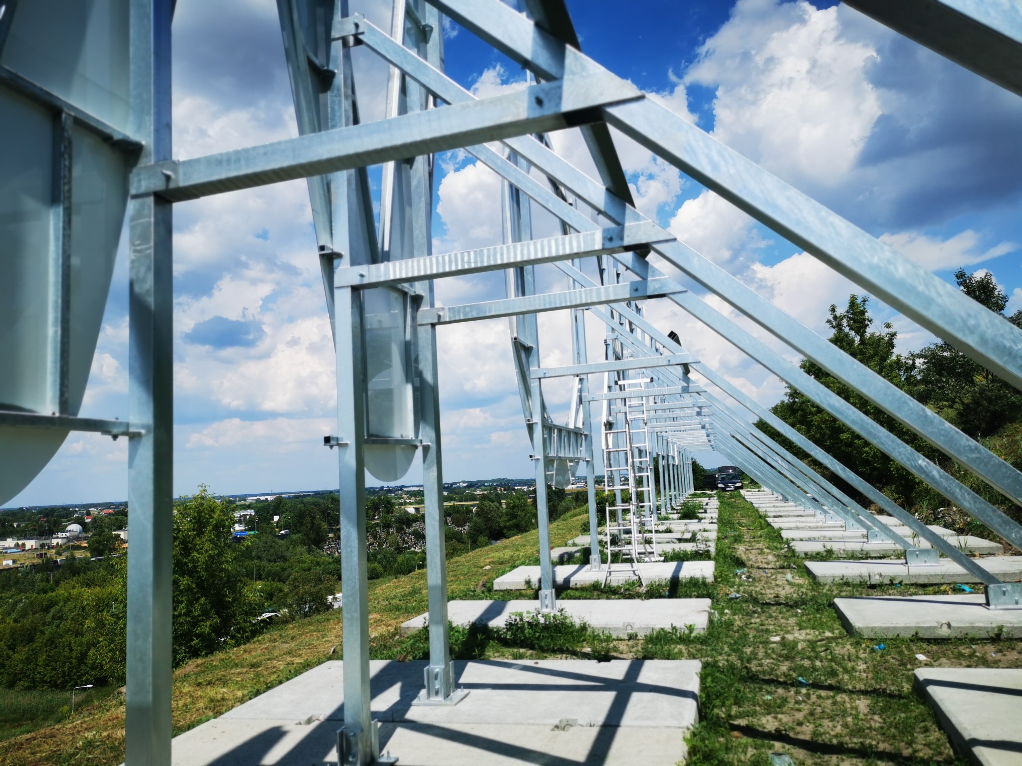 Metalowa konstrukcja wsporcza z drabinką, prawdopodobnie pod panele słoneczne, na betonowych fundamentach, widok z dołu, w tle zielony krajobraz i błękitne niebo z chmurami.