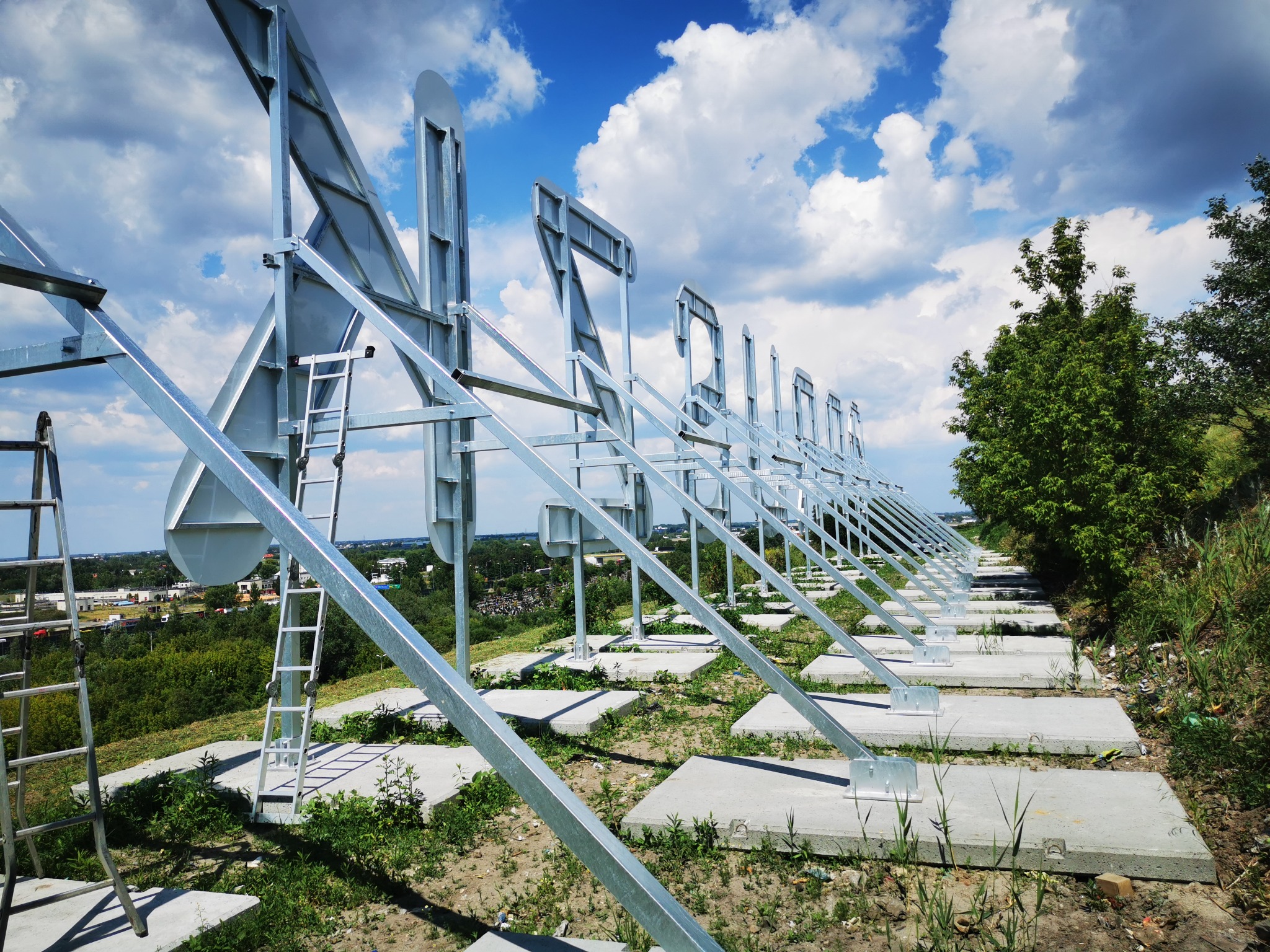 Konstrukcja metalowa z równomiernie rozstawionymi elementami w kształcie litery A, wsparta na betonowych fundamentach, z drabiną opartą o jedną z podpór, na tle zieleni i błękitnego nieba z chmurami.