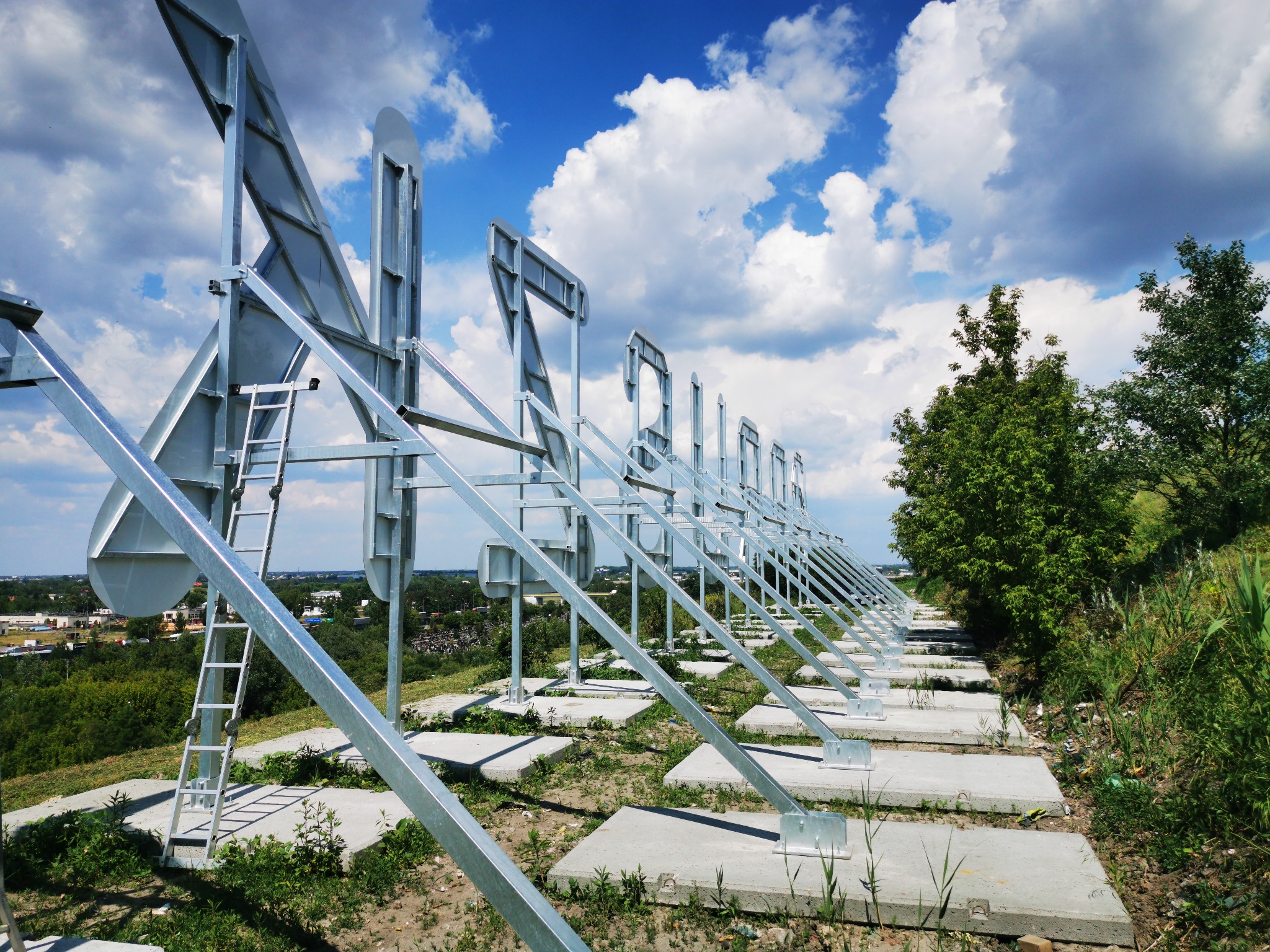 Konstrukcja metalowa z drabinką na betonowych fundamentach na wzgórzu, widok na miasto w tle pod błękitnym niebem z chmurami.