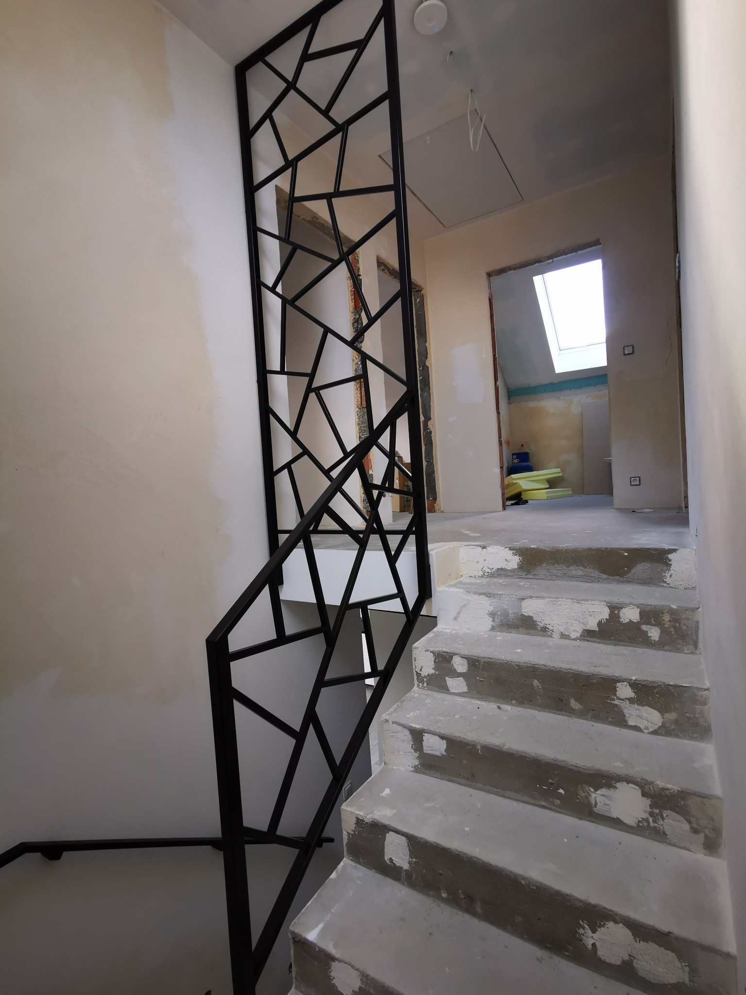 Metalowa balustrada schodowa w stylu geometrycznym, czarna, zamontowana przy surowych schodach betonowych w trakcie wykończenia wnętrza domu.