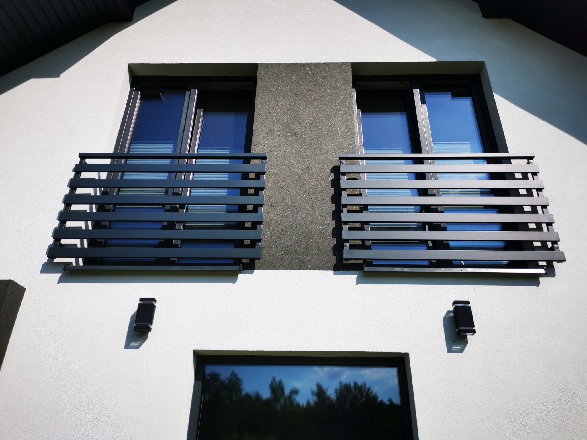 Dwa balkony z nowoczesnymi, poziomymi balustradami w kolorze grafitowym, zamontowane przy oknach na białej fasadzie domu z szarą wstawką. Widoczne czarne lampy zewnętrzne i odbicie drzew w dolnym...